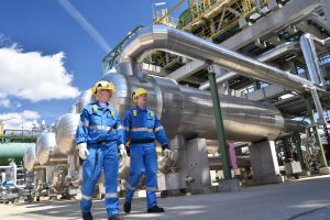 Segurança em atmosferas explosivas: Dois homens de meia-idade vestindo macacões azuis e capacetes de segurança em frente a uma instalação industrial com grandes tubos de metal e equipamentos.