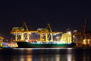 Setor naval: Um grande navio de carga atracado em um porto à noite, com grandes guindastes e edifícios iluminados ao fundo