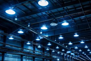 Luminárias LED: Teto industrial com várias luzes azuis penduradas em vigas de metal, criando uma atmosfera brilhante e moderna. O fundo apresenta uma estrutura metálica típica de um armazém ou ambiente de fábrica.